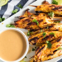 grilled chicken satay on a white platter with peanut sauce and a wedge of lime