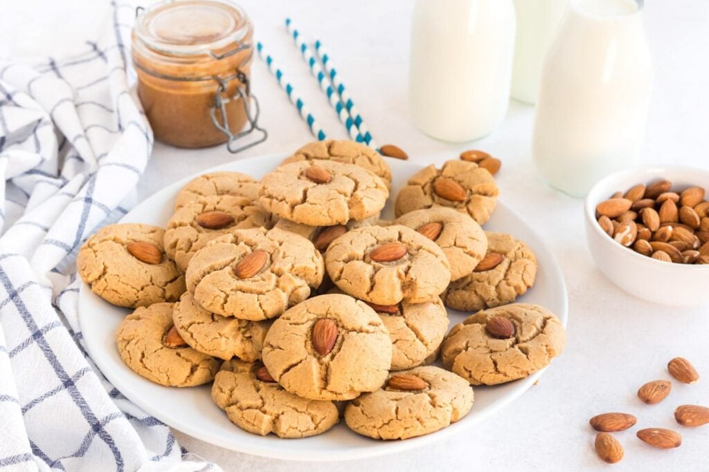 almond butter cookies on a white plate with almonds, milk an d a jar of almond butter
