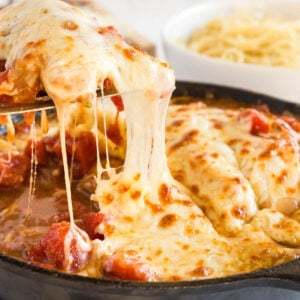 A skillet of mozzarella chicken parmesan topped with melted, stretchy cheese and tomato sauce, with a serving being lifted out; a bowl of spaghetti is in the background.