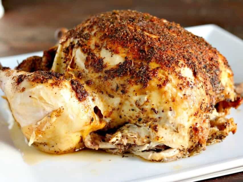 A whole roasted chicken, seasoned with a blend of herbs and spices, sits on a white plate. The skin is crispy and golden brown, with some juices visible at the bottom of the plate.
