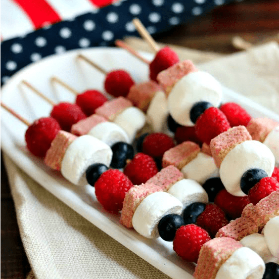 red white and blue fruit and cookie kabobs on a white plate with an american flag in the background