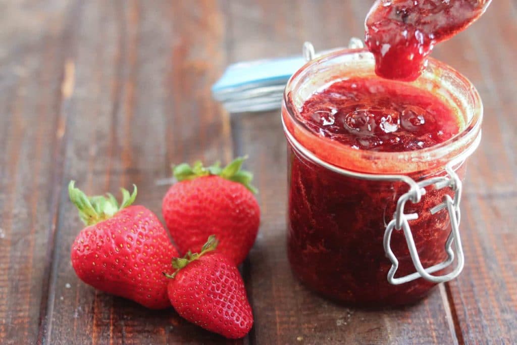jar of strawberry jam with a spoon showing texture with strawberries alongside it