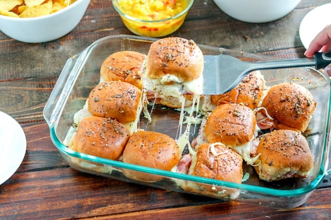 Spatula holding ham and pineapple slider with remaining sliders in glass baking dish beneath on wooden table.