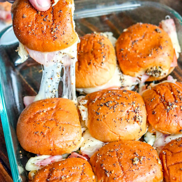 Pulling a hot, melty ham and pineapple slider out of glass baking dish with remaining sliders in it.
