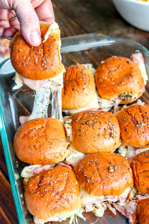Pulling a hot, melty ham and pineapple slider out of glass baking dish with remaining sliders in it.