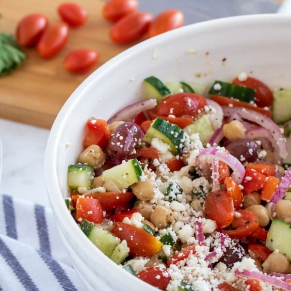 greek chick pea salad in a white bowl with cucumbers and tomatoes on a chopping board.