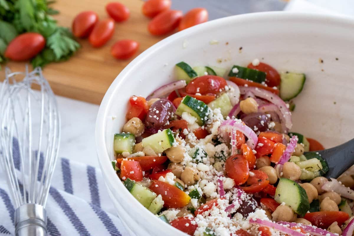 greek chick pea salad in a white bowl with cucumbers and tomatoes on a chopping board.
