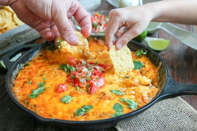 skillet of queso dip with 2 hands using chips to dip