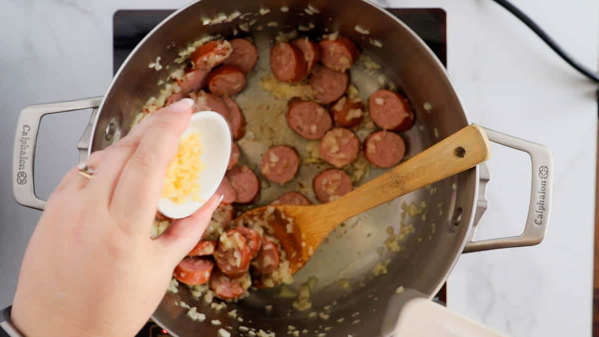a skillet with sliced sausage in it, and a hand adding garlic.