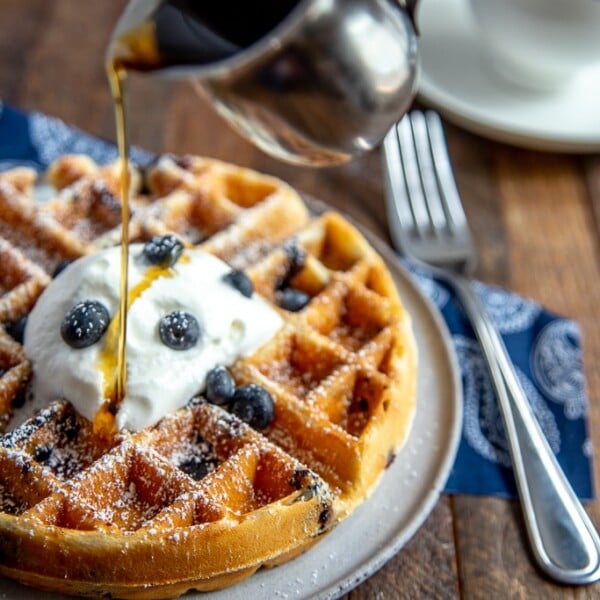Blueberry waffle with powdered sugar, whipped cream and blueberries with syrup being poured over the top