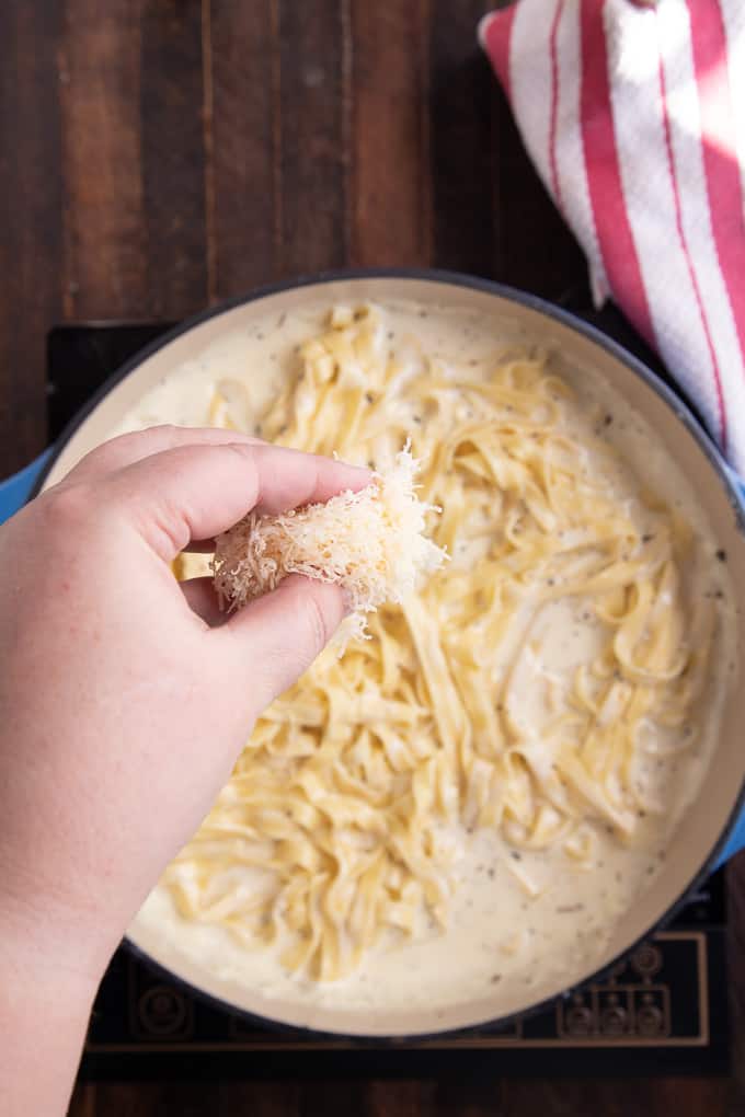 skillet with fettuccine alfredo with a hand holding parmesan.