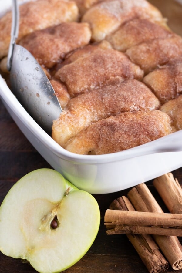 apple dumplings in a white casserole dish with a cut apple and cinnamon sticks