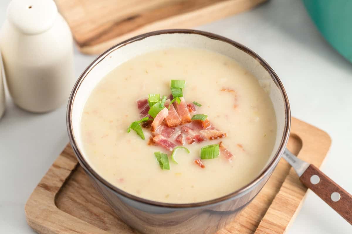 A bowl of creamy leek and potato soup, garnished with chopped bacon and green onions, sits on a wooden tray with a spoon beside it.