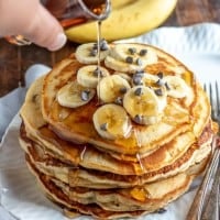 stack of banana pancakes on a plate with syrup being poured over the top.