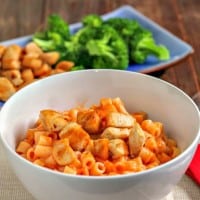 Chicken tomato pasta in a white bowl with broccoli and chicken in the background.