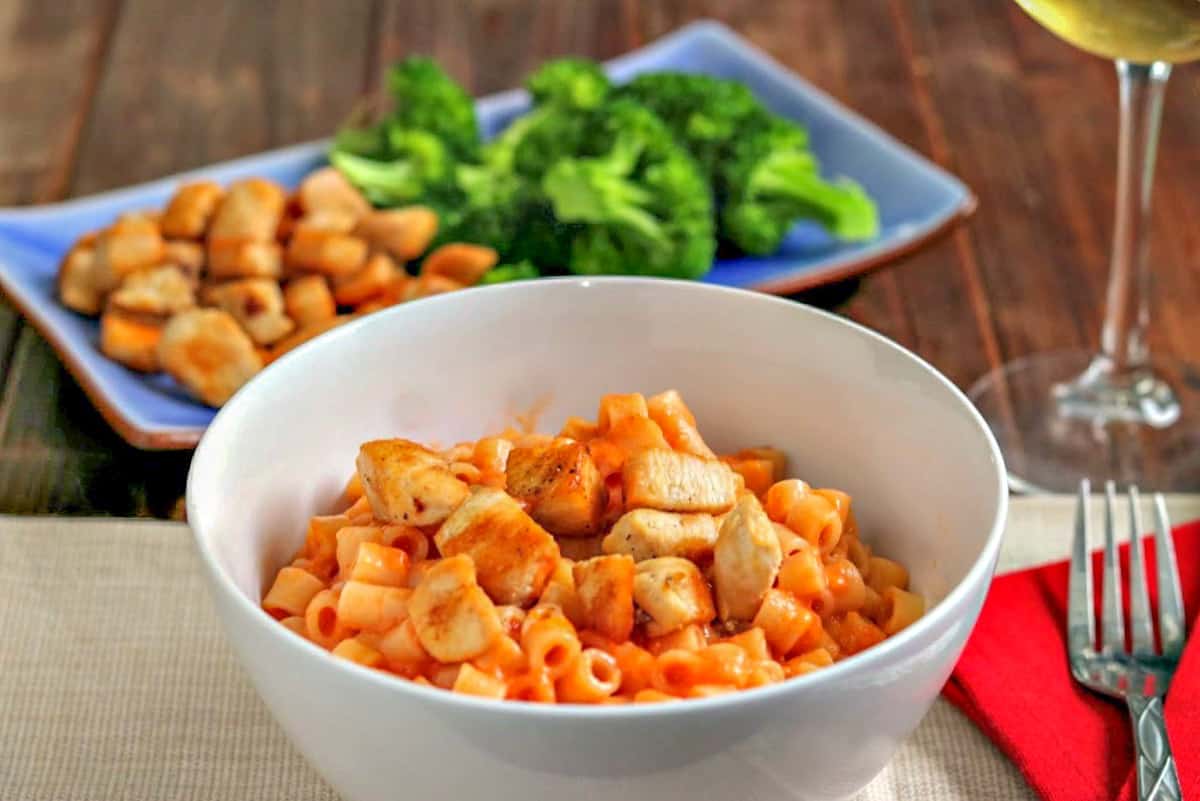 Chicken tomato pasta in a white bowl with broccoli and chicken in the background.