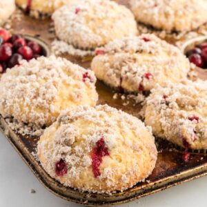 A metal muffin pan filled with cranberry muffins topped with crumbly streusel, some fresh cranberries visible among the warm, golden muffins.