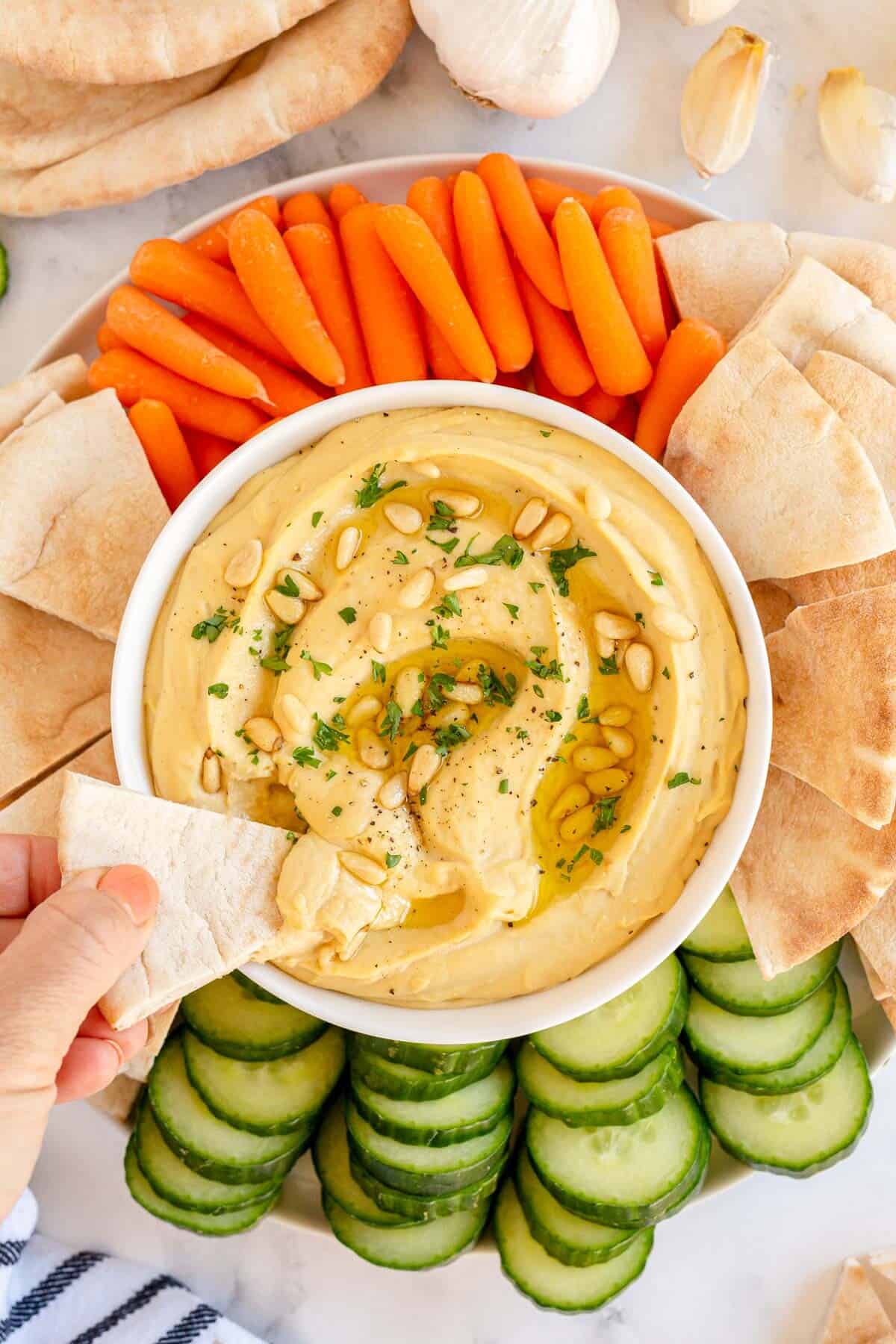 roasted garlic hummus in a white bowl with pita bread, carrots and cucumbers with a hand and pita bread