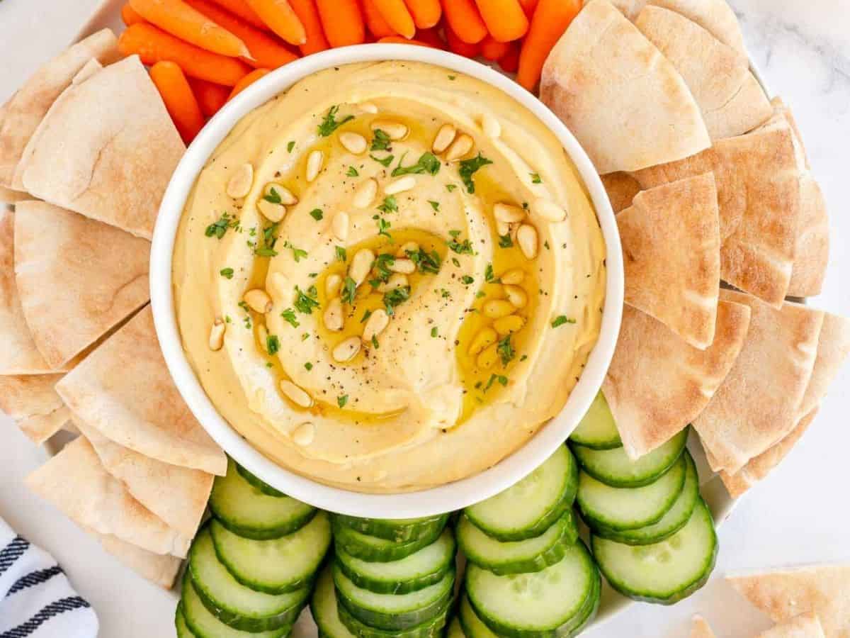 roasted garlic hummus in a white bowl with pita bread, carrots and cucumbers