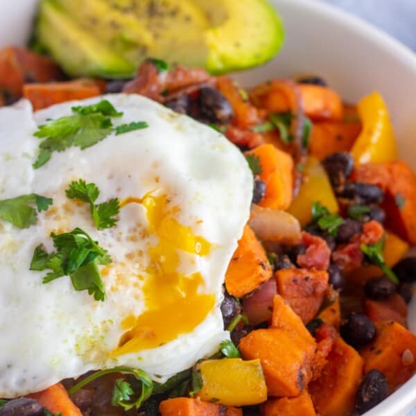 sweet potato hash with an egg, and avocado slices in a white bowl