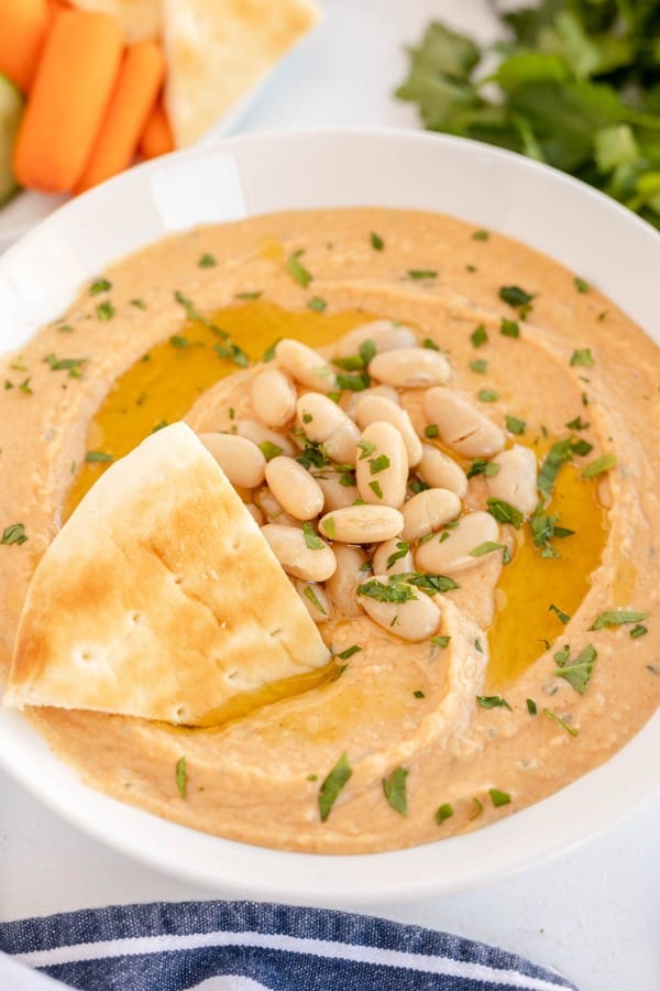 a white bowl of white bean hummus with a wedge of pita bread in it.