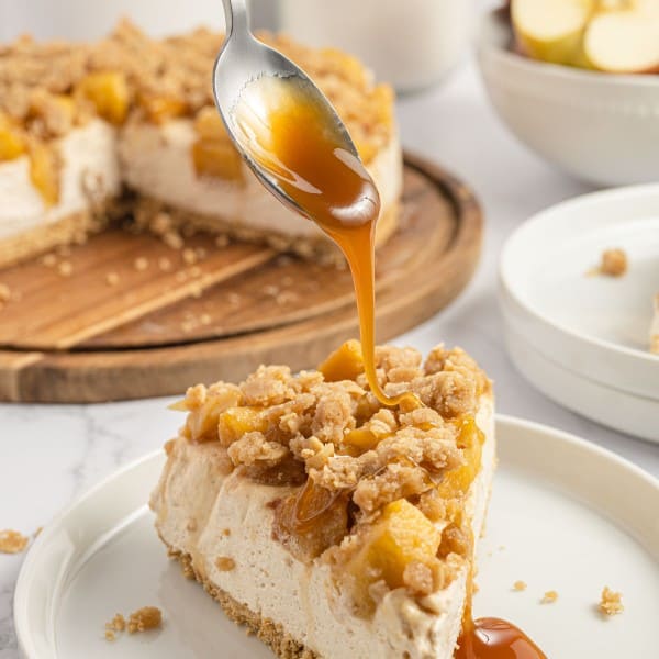slice of apple crisp cheesecake on a white plate with caramel being poured over the top.