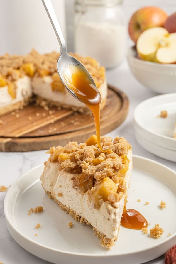 slice of apple crisp cheesecake on a white plate with caramel being poured over the top.