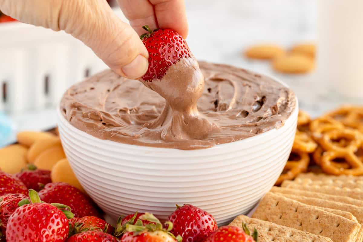 A person is dipping a strawberry into a bowl of Brownie Batter Dip.