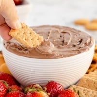 A person is dipping a graham cracker into a bowl of Brownie Batter Dip.