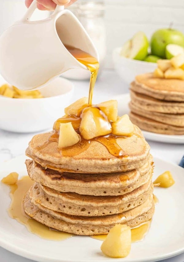 Pouring syrup onto a stack of pancakes with apples.