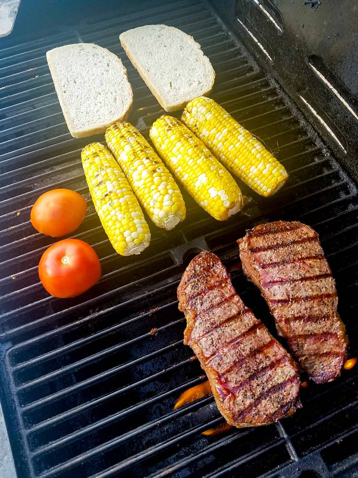 cooked steaks on a grill with tomatoes, corn and bread.