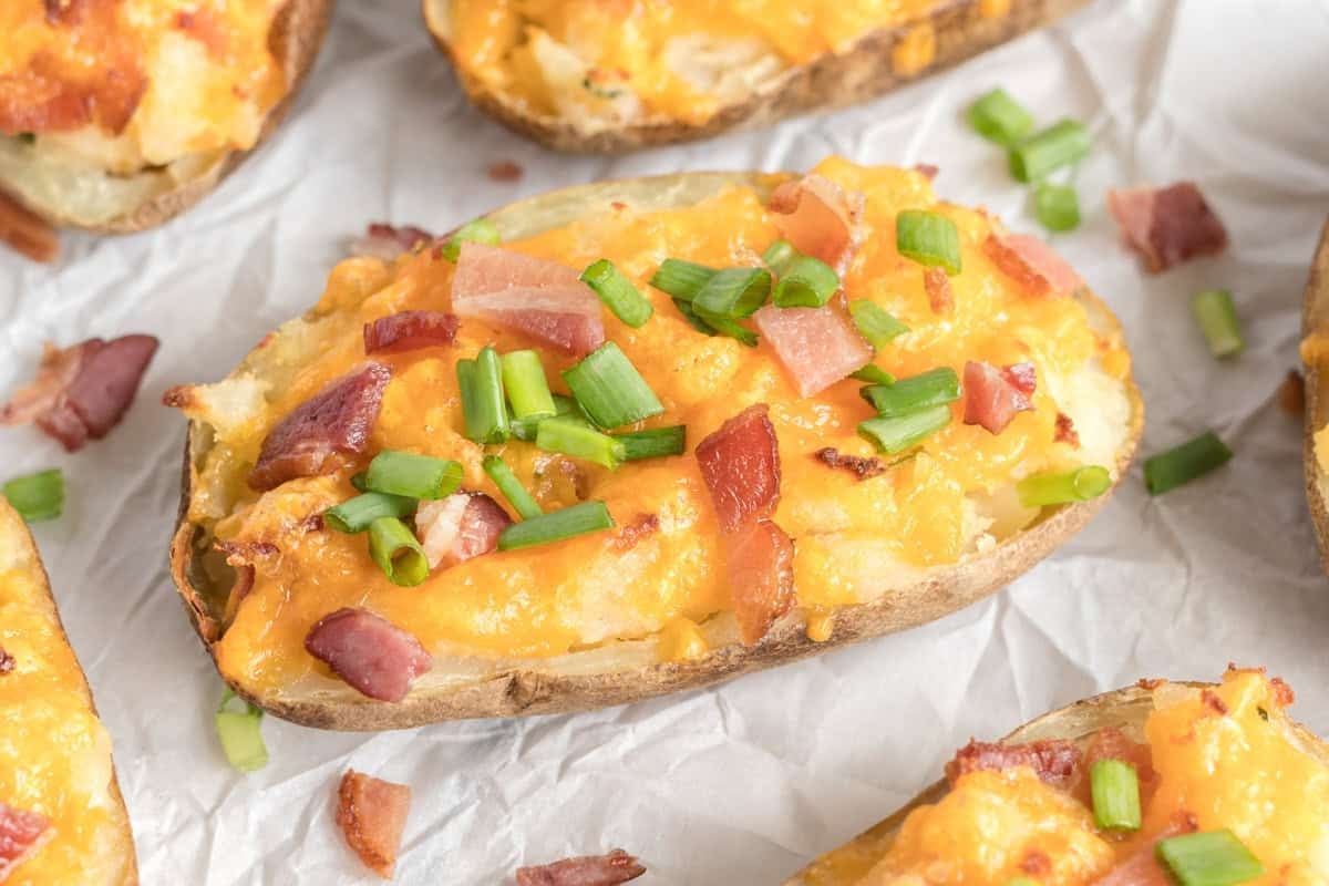A loaded twice baked potato topped with melted cheddar cheese, chopped bacon, and green onions on parchment paper.