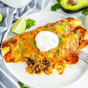 A plate with two enchiladas topped with melted cheese, sour cream, and cilantro. A fork is on the plate, with a halved avocado, a lime wedge, and a blue-striped cloth in the background.