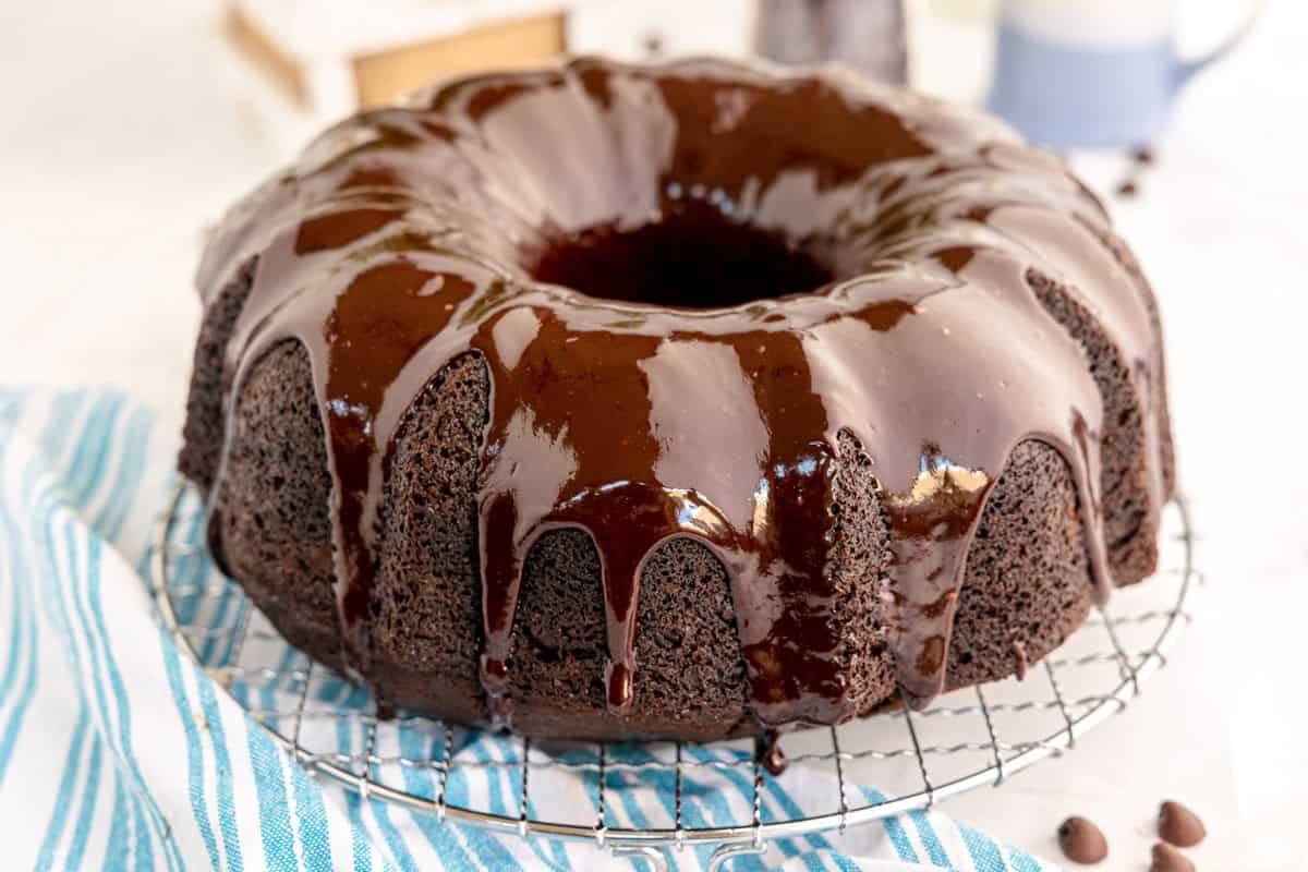 A chocolate bundt cake with glossy chocolate glaze sits on a wire rack over a striped towel.