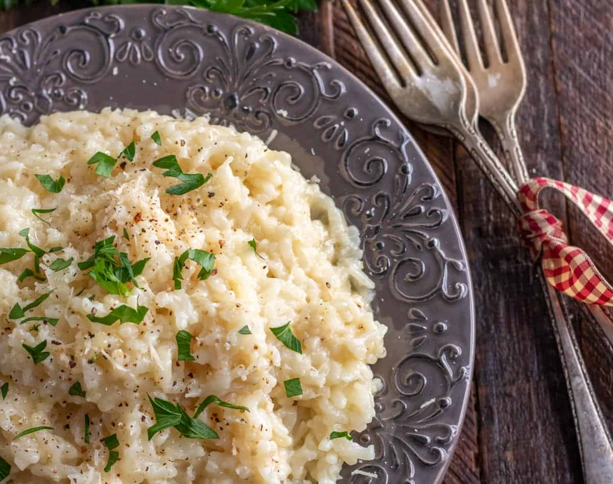 A plate of creamy risotto topped with chopped parsley and black pepper, served on a decorative plate with two forks beside it on a wooden table.