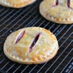 A round hand pie with golden, flaky crust and two slits showing red fruit filling, cooling on a black wire rack.