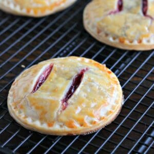 A round hand pie with golden, flaky crust and two slits showing red fruit filling, cooling on a black wire rack.