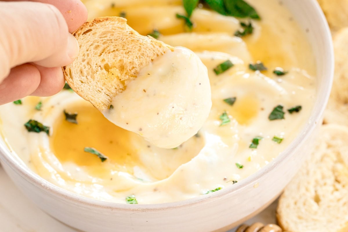 A hand dips a slice of bread into a bowl of creamy cheese dip, garnished with chopped herbs and drizzled with honey.