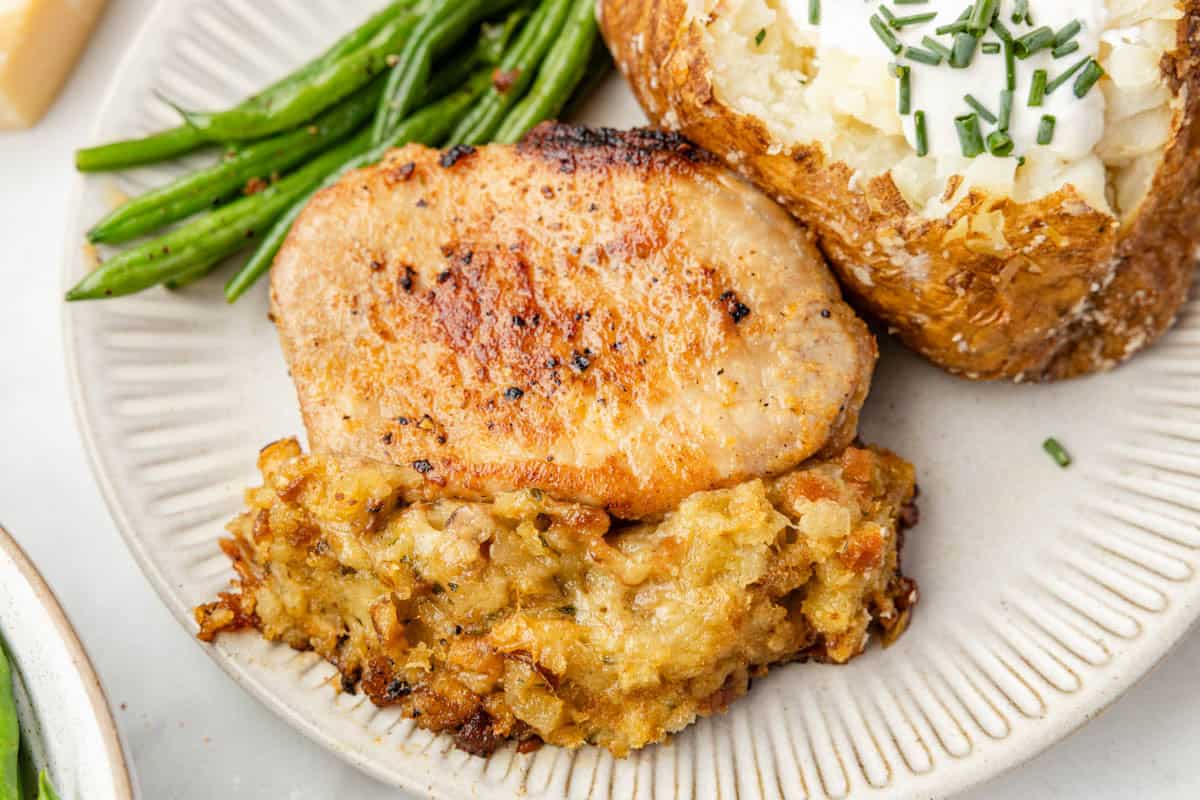 A pork chop with stuffing on a plate next to green beans and a baked potato.