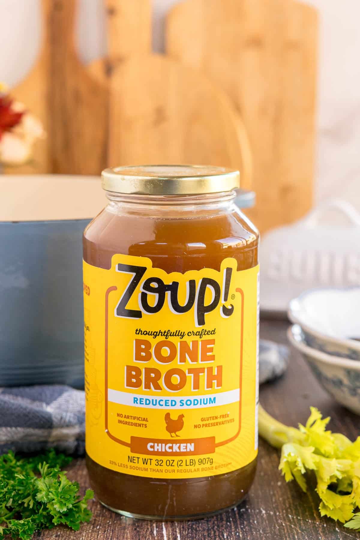 A jar of Zoup! chicken bone broth on a table with fresh herbs and celery. Text on the image says 'Bone Broth'.