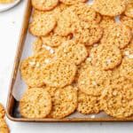 Lace cookies on a parchment-lined baking sheet with oats sprinkled around.