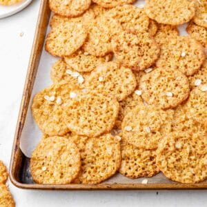 Lace cookies on a parchment-lined baking sheet with oats sprinkled around.