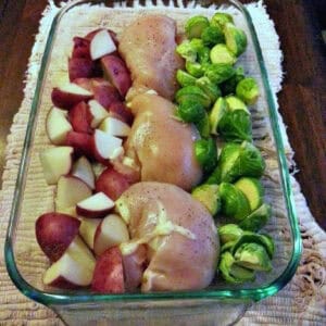 Glass baking dish containing raw chicken breasts, halved Brussels sprouts, and chopped red potatoes, all arranged in separate rows, ready for cooking.