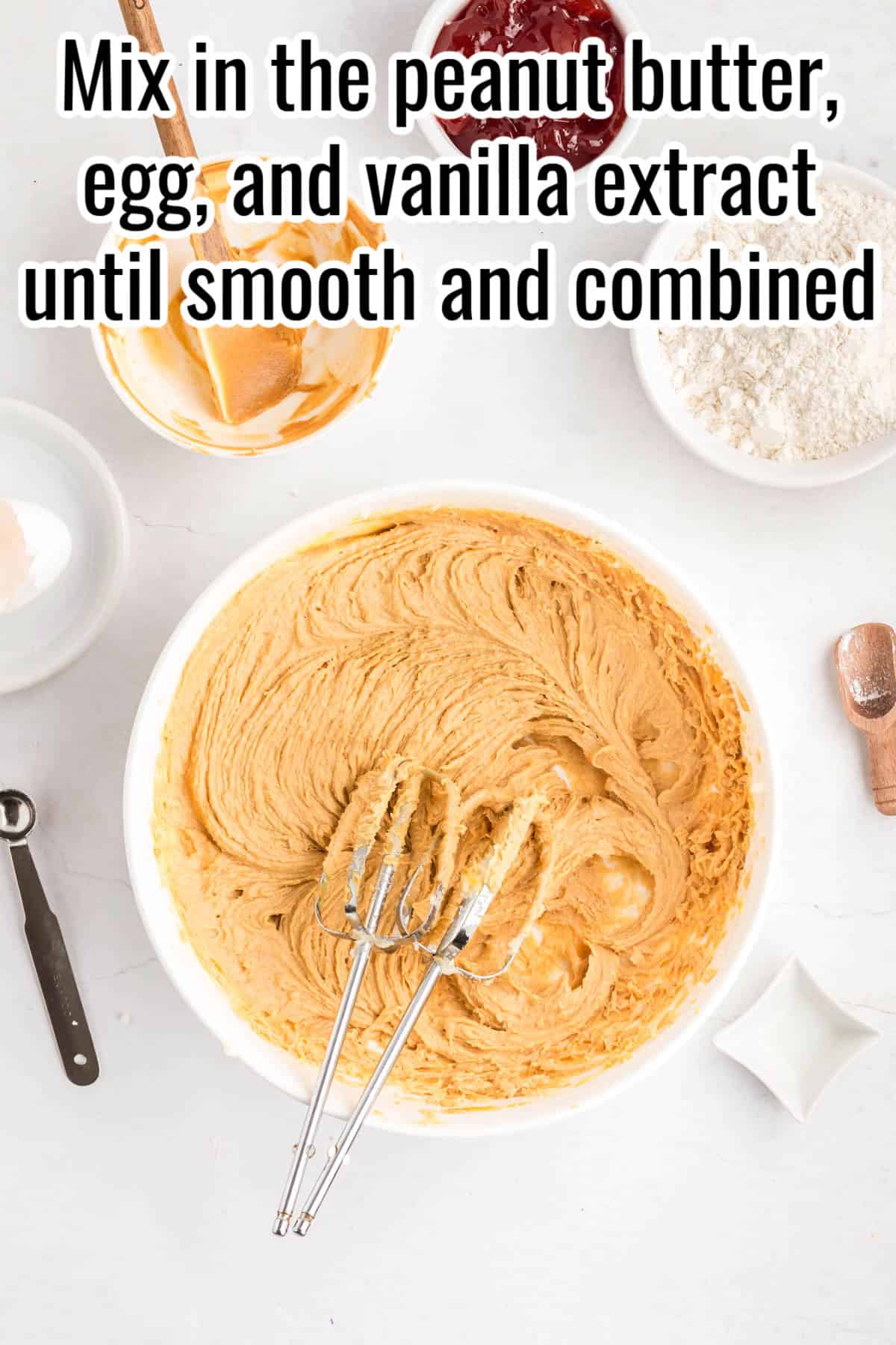 A bowl of peanut butter batter with beaters inside, surrounded by ingredients. Text on the image says 'Mix in the peanut butter, egg, and vanilla extract until smooth and combined'.