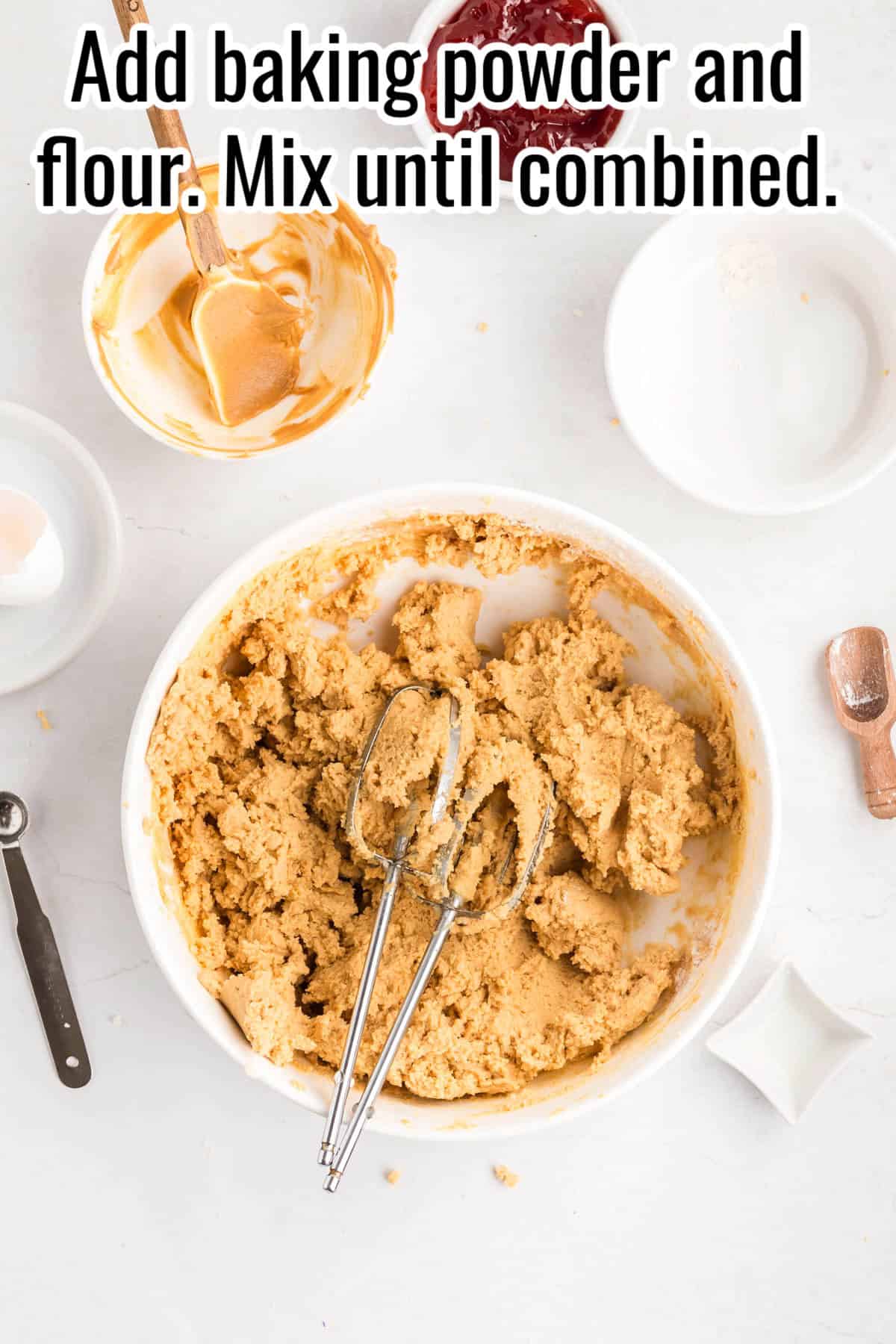 A mixing bowl with thick peanut butter dough and beaters. Text on the image says 'Add baking powder and flour. Mix until combined'.