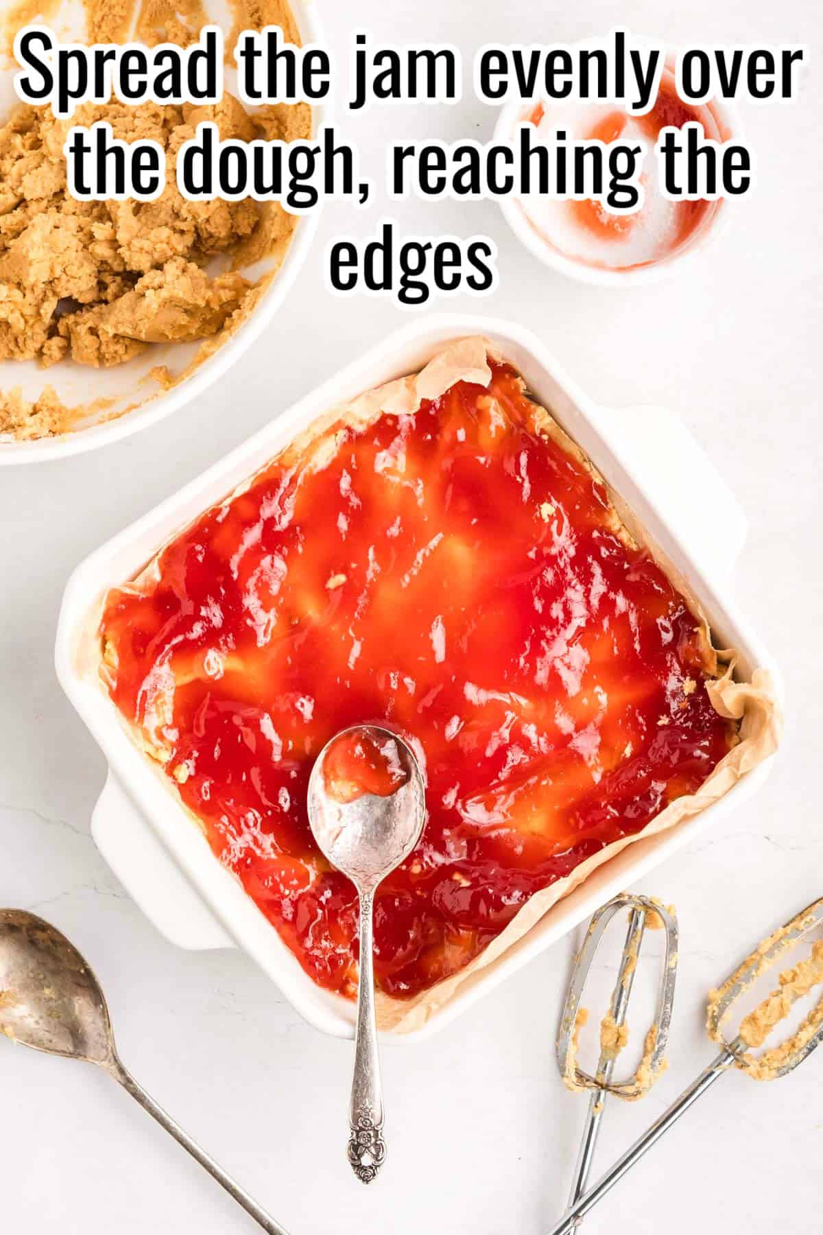 A baking dish filled with dough spread with red jam. Text on the image says 'Spread the jam evenly over the dough, reaching the edges'.