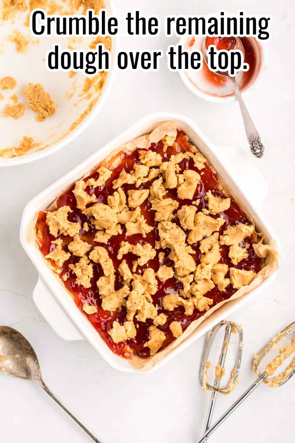 A baking dish of jam-topped dough with crumbled dough added on top. Text on the image says 'Crumble the remaining dough over the top'.