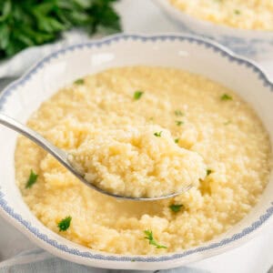 Close-up of a spoonful of pastina above a bowl, with small parsley leaves scattered on top.