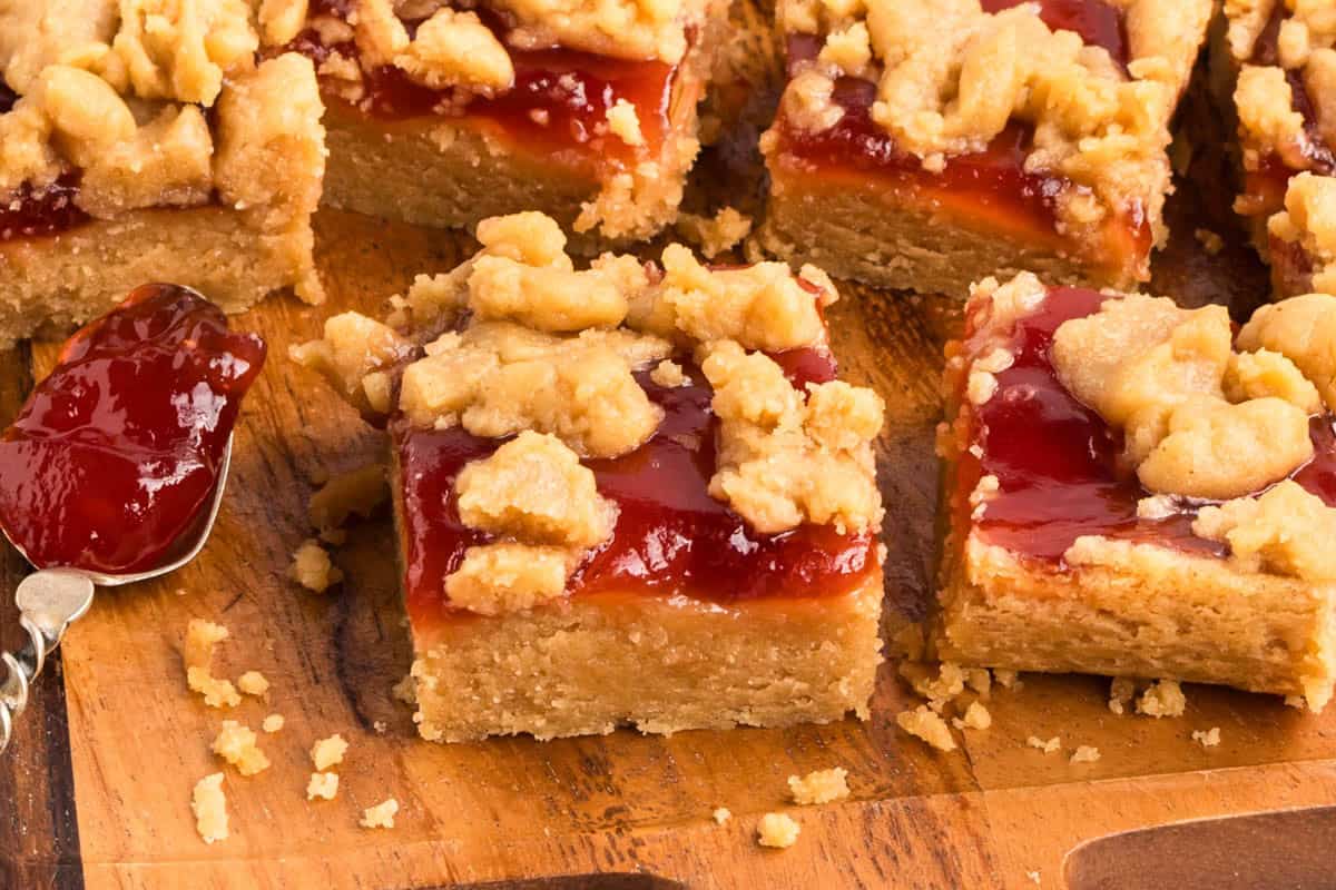 Several peanut butter jam bars on a wooden board with a spoonful of jam nearby.