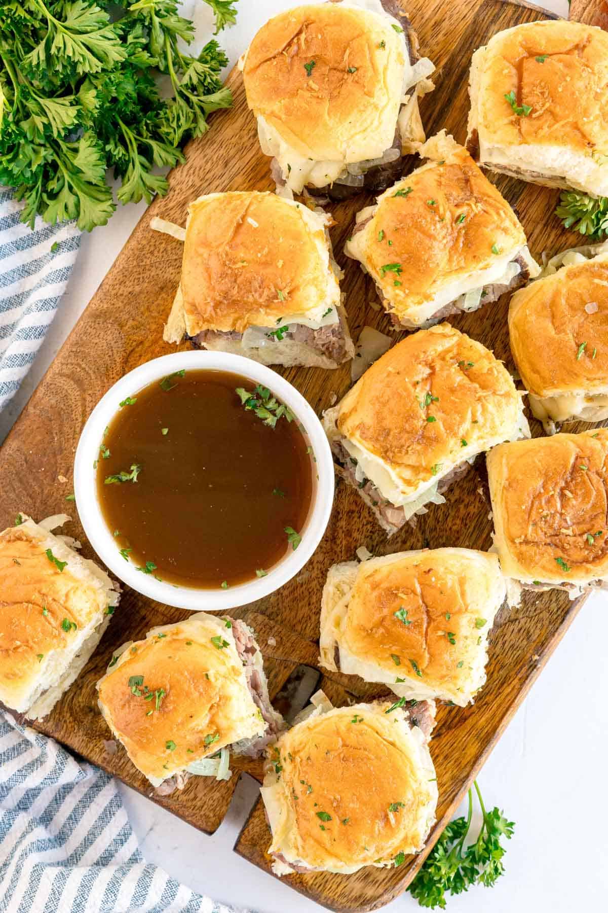 Overhead view of multiple roast beef sliders and a bowl of au jus on a wooden board.
